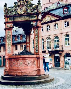 Isabell Rahms betrachtet die Inschriften am Mainzer Marktbrunnen von 1526 aus rotem Sandstein, im Hintergrund ist der Mainzer Dom zu sehen.