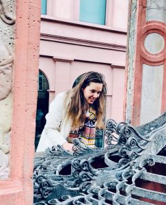 Isabell Rahms lehnt sich über das kunstvolle Schmiedegitter des Mainzer Marktbrunnens und blickt in den historischen Brunnen.