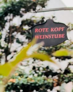Schwarzes Schild mit der roten Aufschrift „Rote Kopf Weinstube“ vor dunklem Hintergrund, im Vordergrund Pflanzen aus der Rotekopfgasse in Mainz.