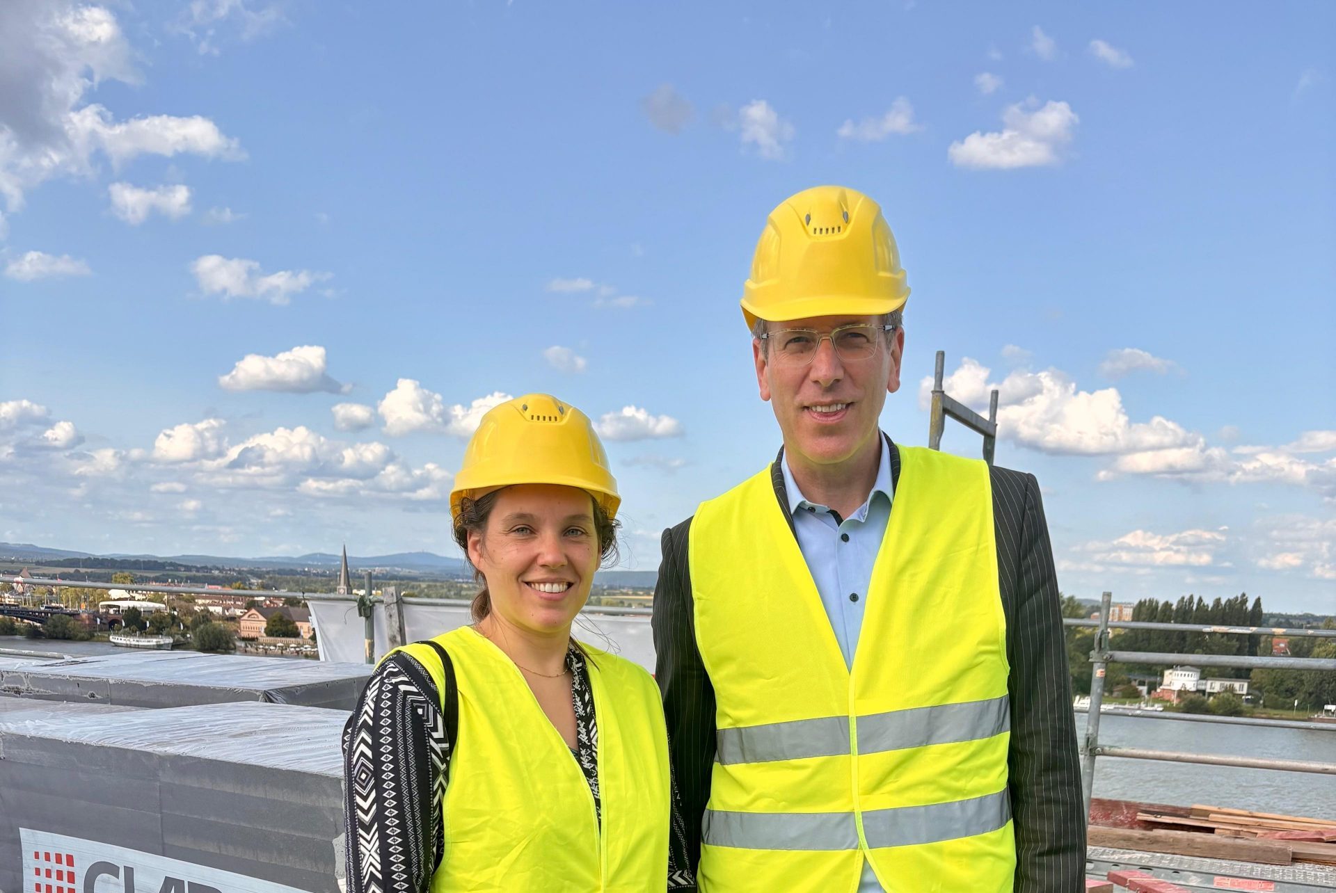 Stadträtin Isabell Rahms und Dezernent Ludwig Holle auf dem Dach der Baustelle des Mainzer Rathauses mit Blick auf den Rhein, in gelben Warnwesten und Schutzhelmen