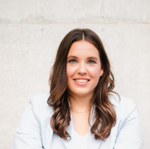 Isabell Rahms, Landtagskandidatin, im hellen Top und hellblauem Blazer, sitzend vor heller Wand, Portraitfoto