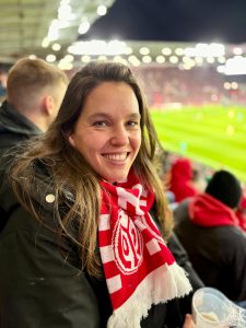 Isabell Rahms, CDU-Landtagskandidatin für Mainz, sitzt in der MEWA ARENA auf der Tribüne. Sie trägt einen rot-weißen Mainz 05 Schal und zeigt ihre Verbundenheit zur Stadt und zum Verein.