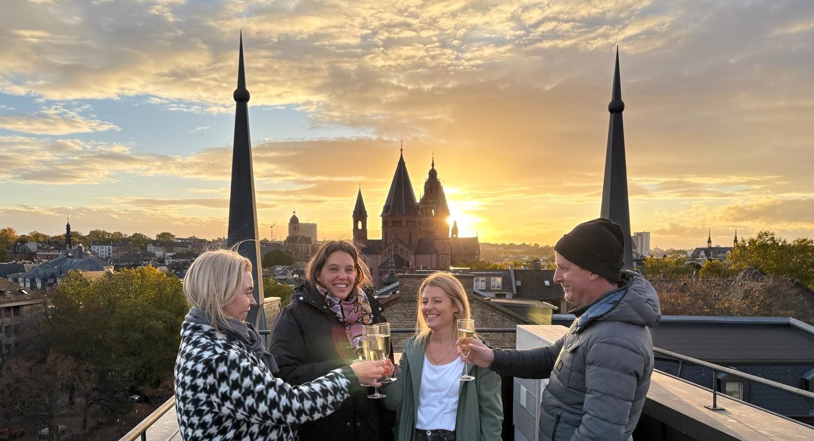 Isabell Rahms mit den Gewinnern der Stadtführung von Isabell Rahms und Merkurist über den Dächern von Mainz. Im Hintergrund der Mainzer Dom bei Sonnenuntergang – Start der Stadtführung mit Begrüßungssekt und Blick auf die Innenstadt Mainz.